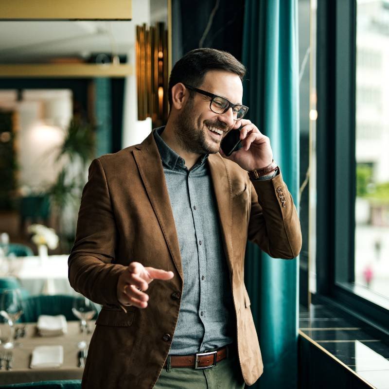 A businessman wearing sun glasses also brown blazer, smiling while talking on his sell phone in a advanced office environment, expressive professional communication and professional success.